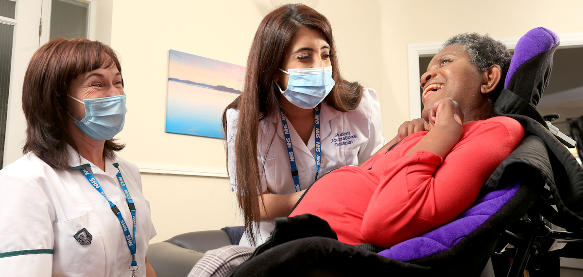 •	Photo of two female healthcare professional wearing masks and smiling at lady with learning disabilities