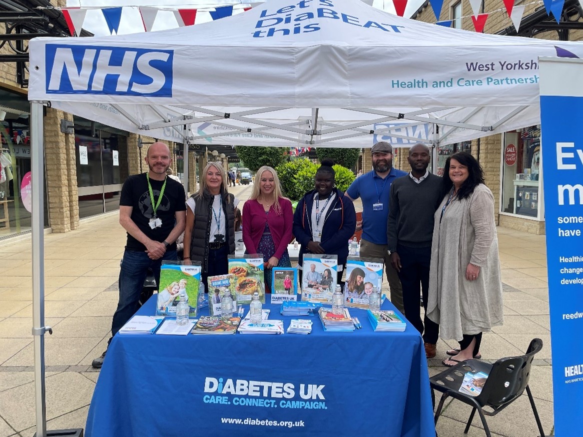 Halifax diabetes stall - a group of men and women standing under a gazebo with 'NHS' on it and behind a table that says 'Diabetes UK' with leaflets on it.