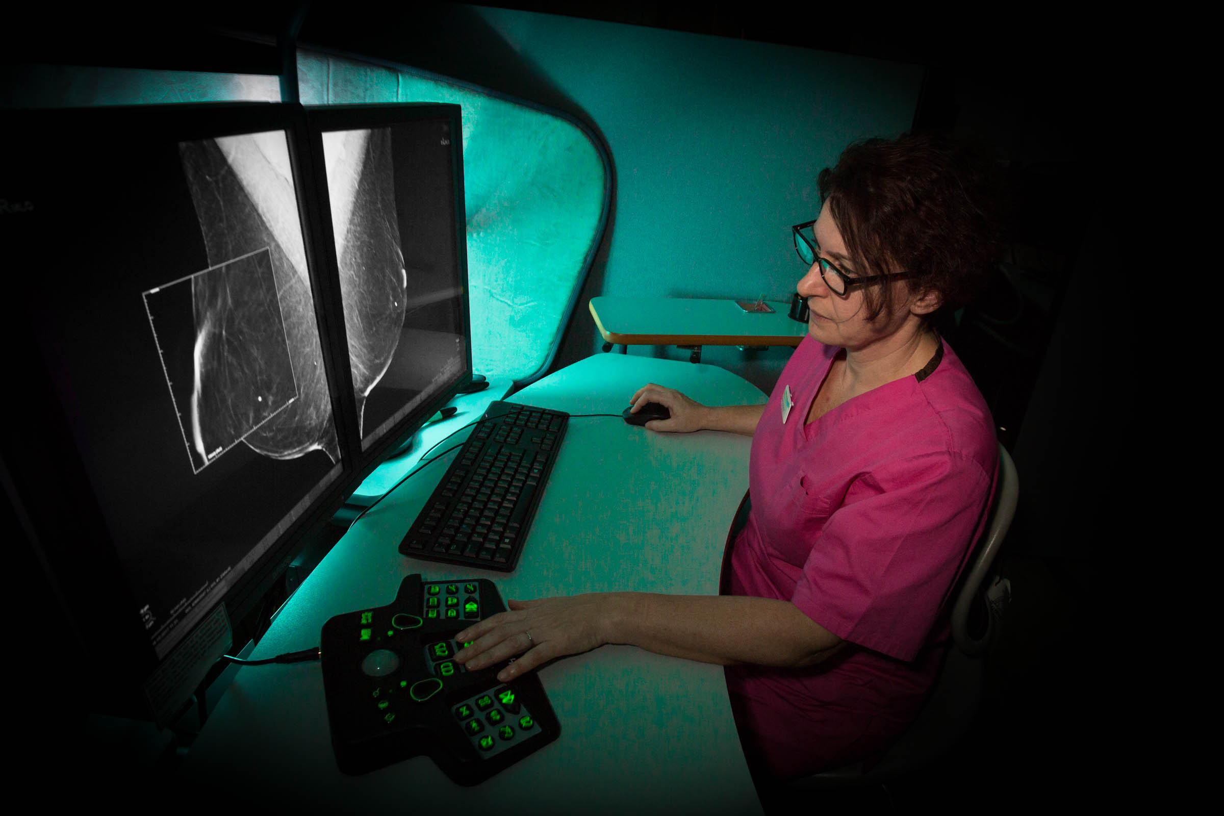 Pennine Breast Screening - technician in a pink uniform siting at a workstation examining scans on a large screen