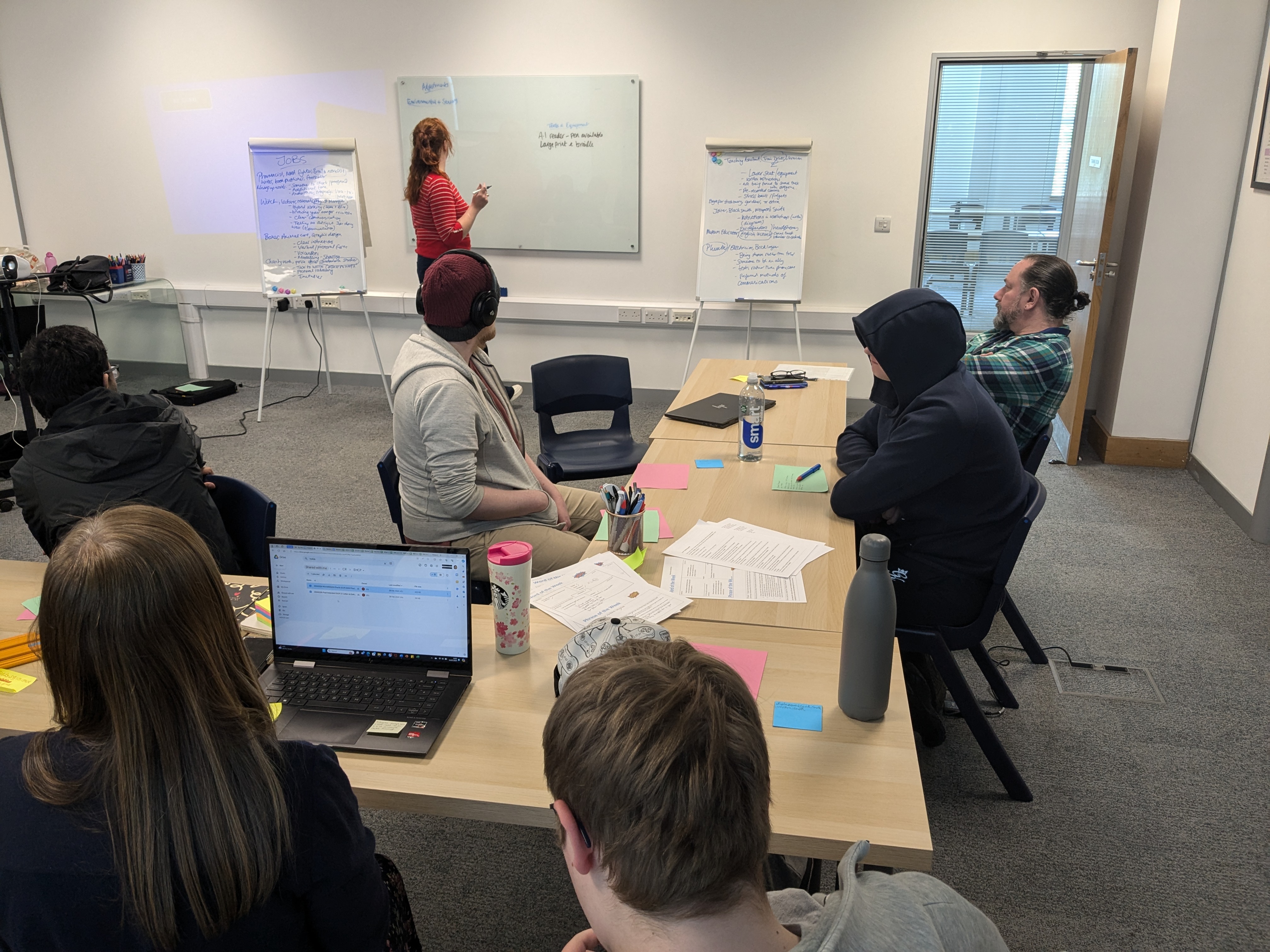 An image of people sat around a desk listening to a woman at the whiteboard.
