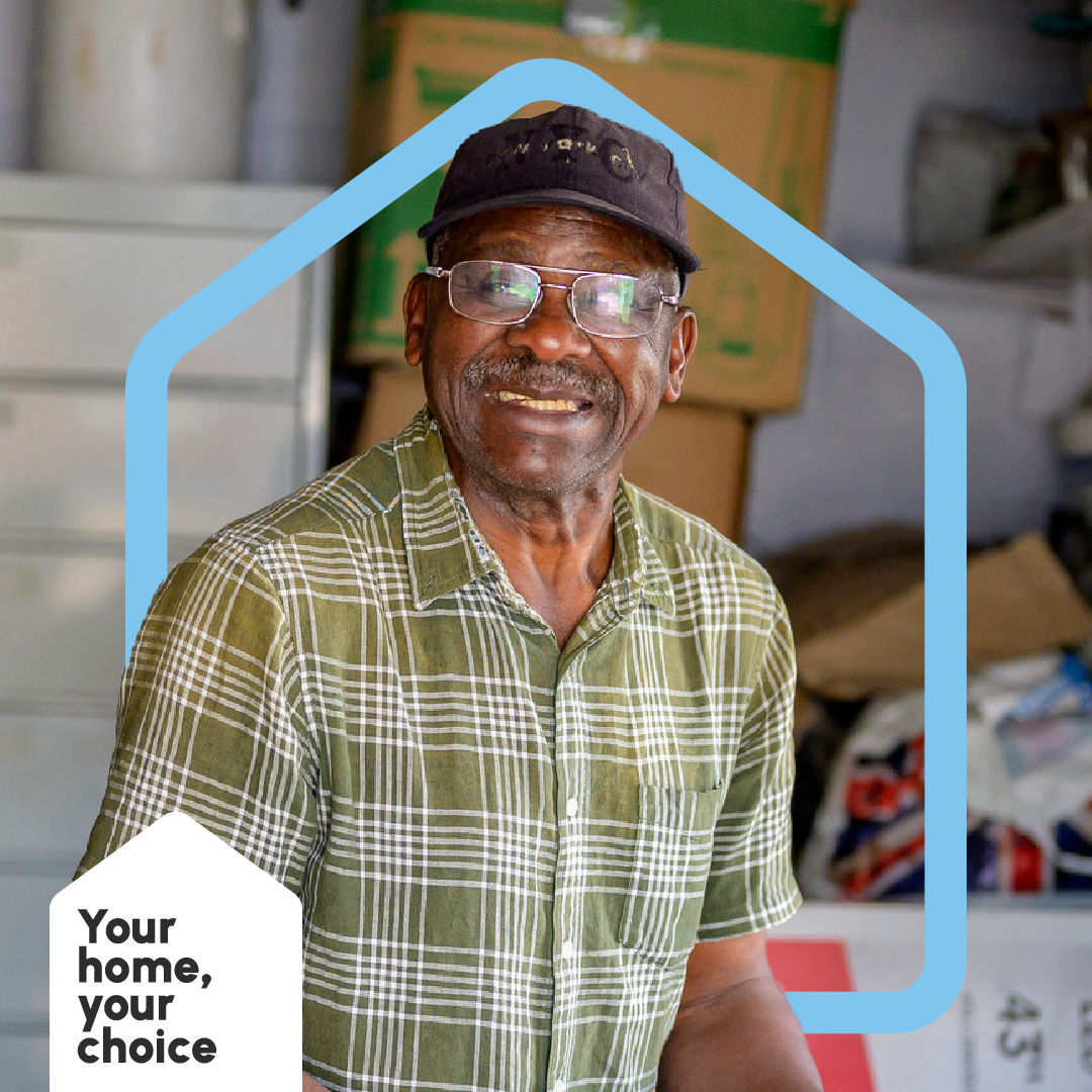 A man standing in a garage, smiling. He is surrounded by a blue illustrated shape of a house and the Your Home, Your Choice logo