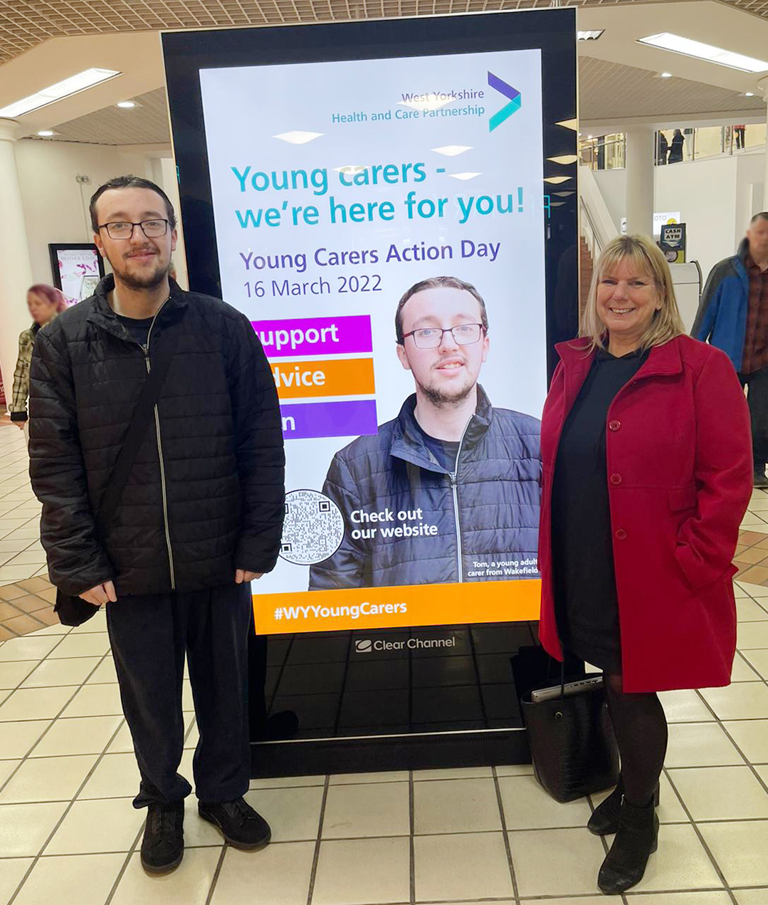 Young adult carer Tom with Young adult carer Tom with Fiona Rogers alongside the Young Carers Action Day billboard poster in The Ridings, Wakefield.