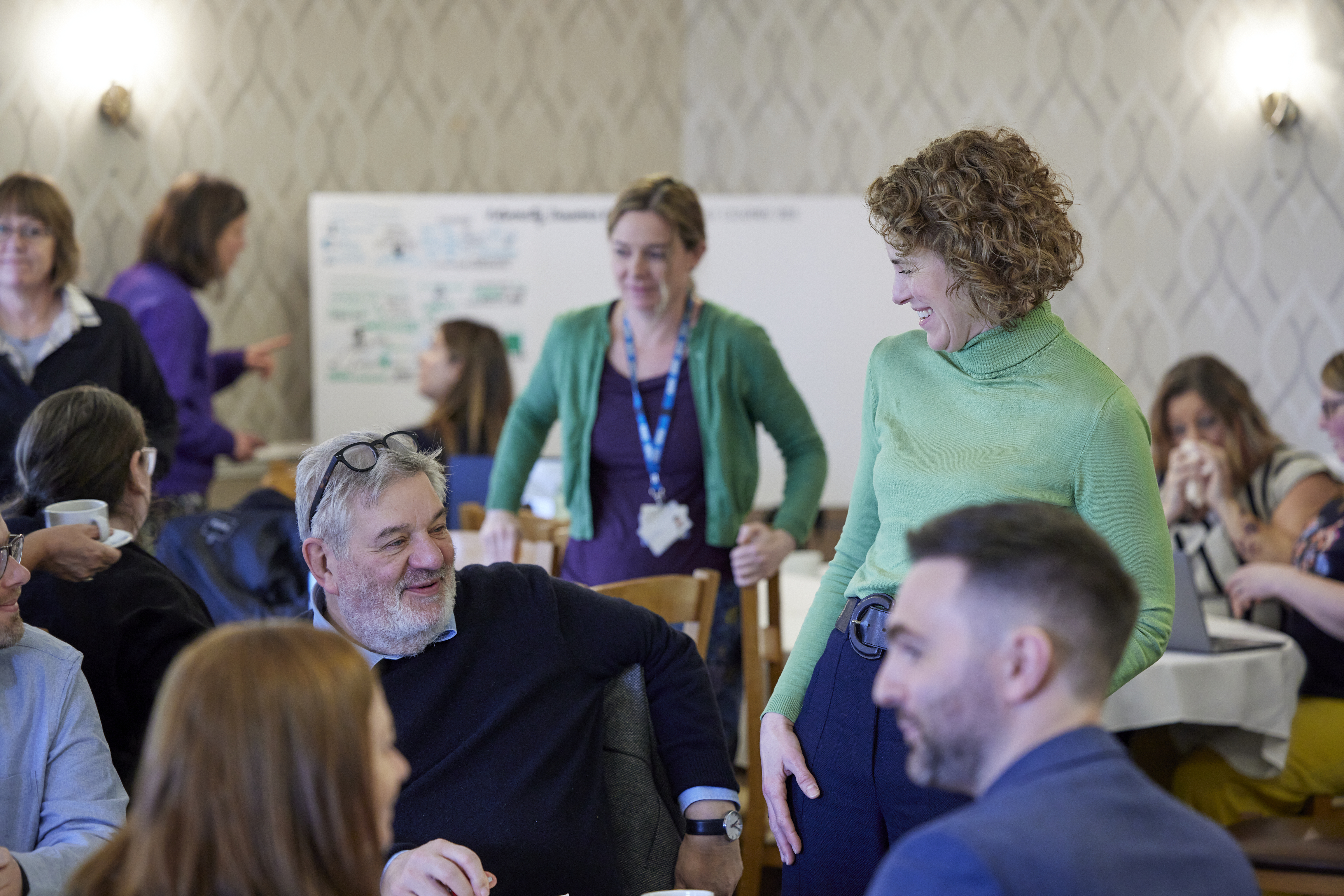 Woman standing in green top smiling down at man in blue jumper, amidst other people at a conference